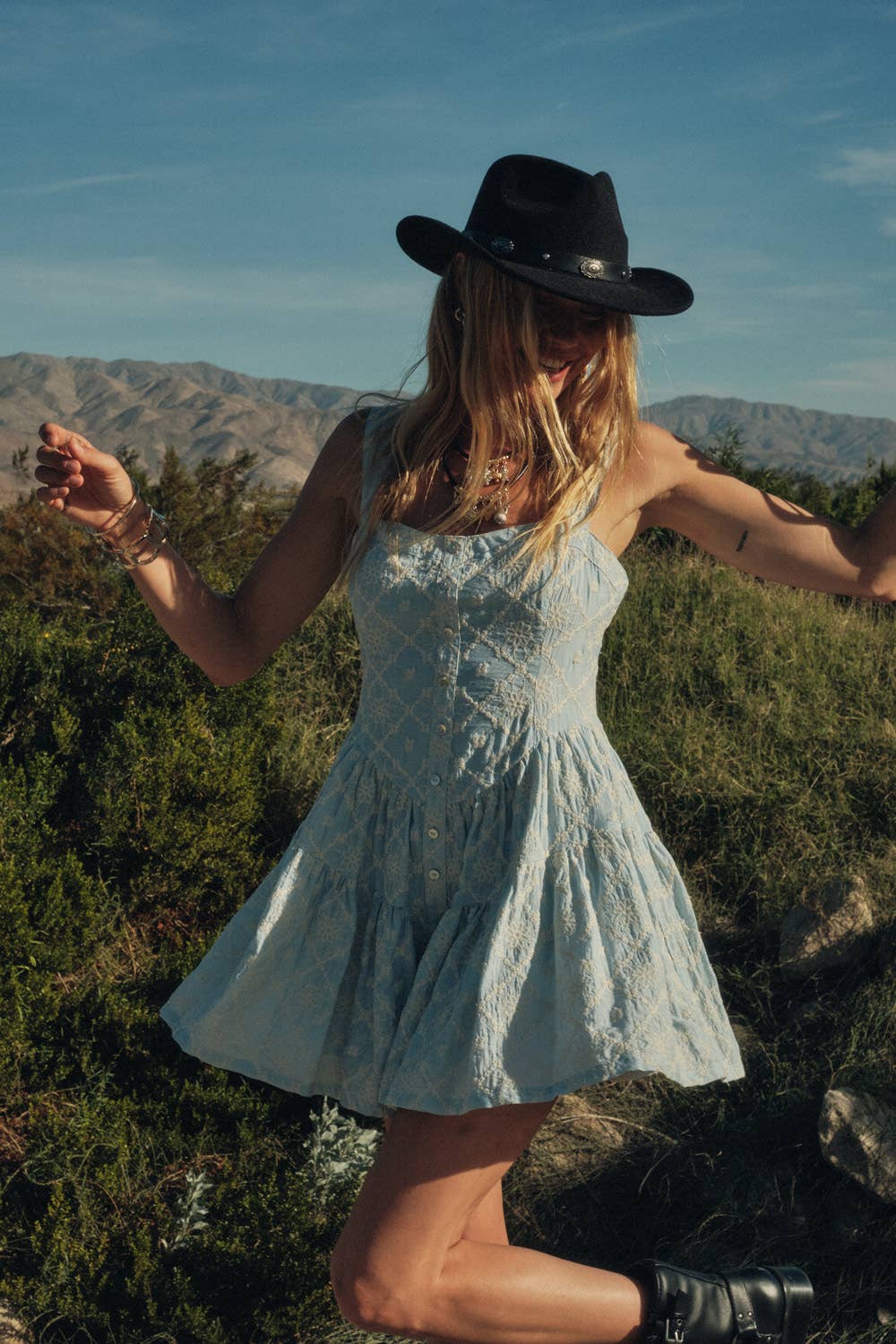 Woman in a light blue dress romper and black hat standing in a natural setting with mountains in the background