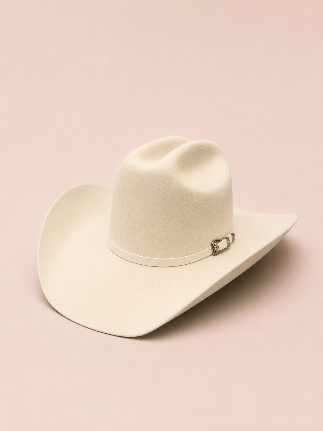 An off-white felt cowboy hat with a silver buckle detail on the band, displayed on a solid pale pink background.
