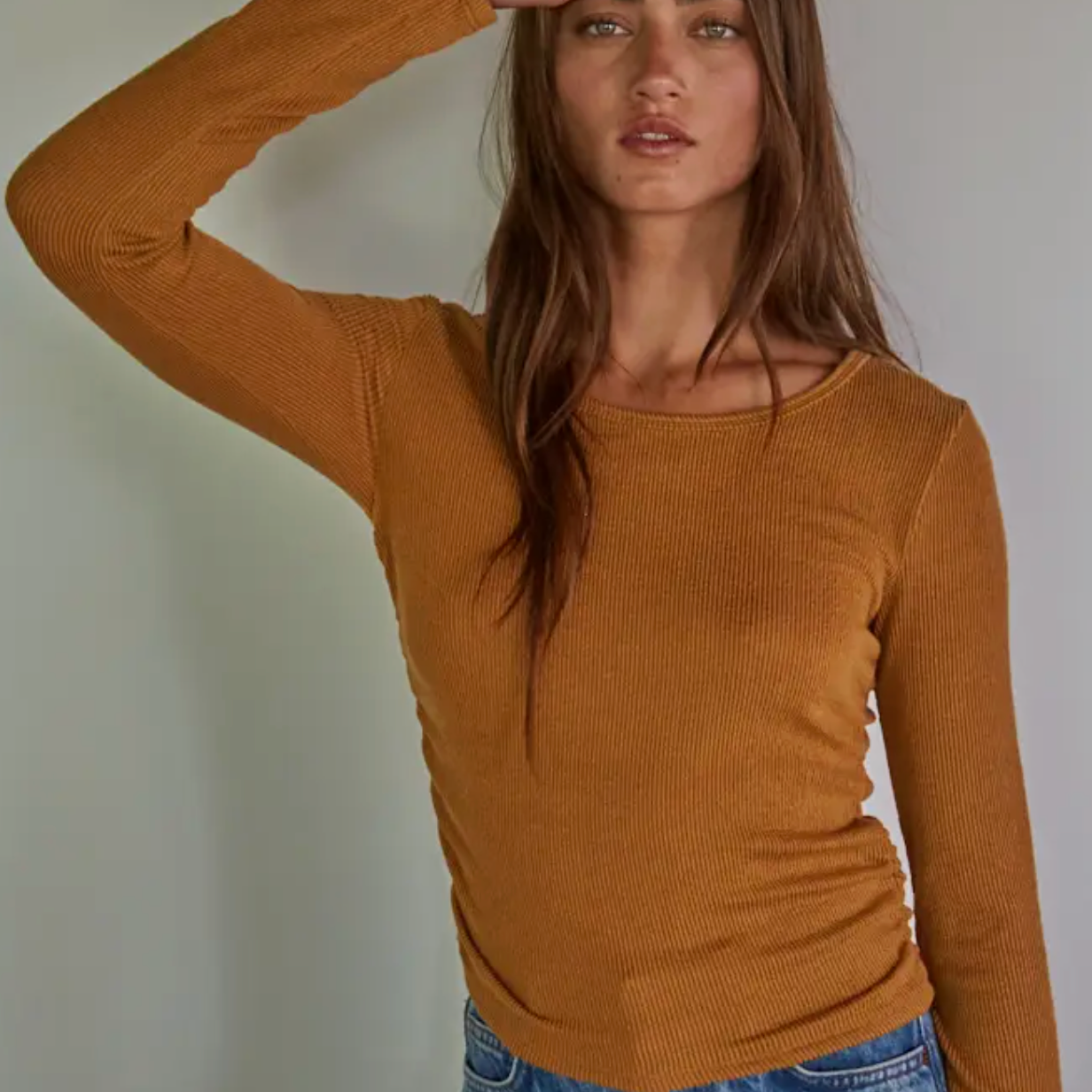 Woman wearing a mustard yellow long-sleeve top and blue jeans against a plain background