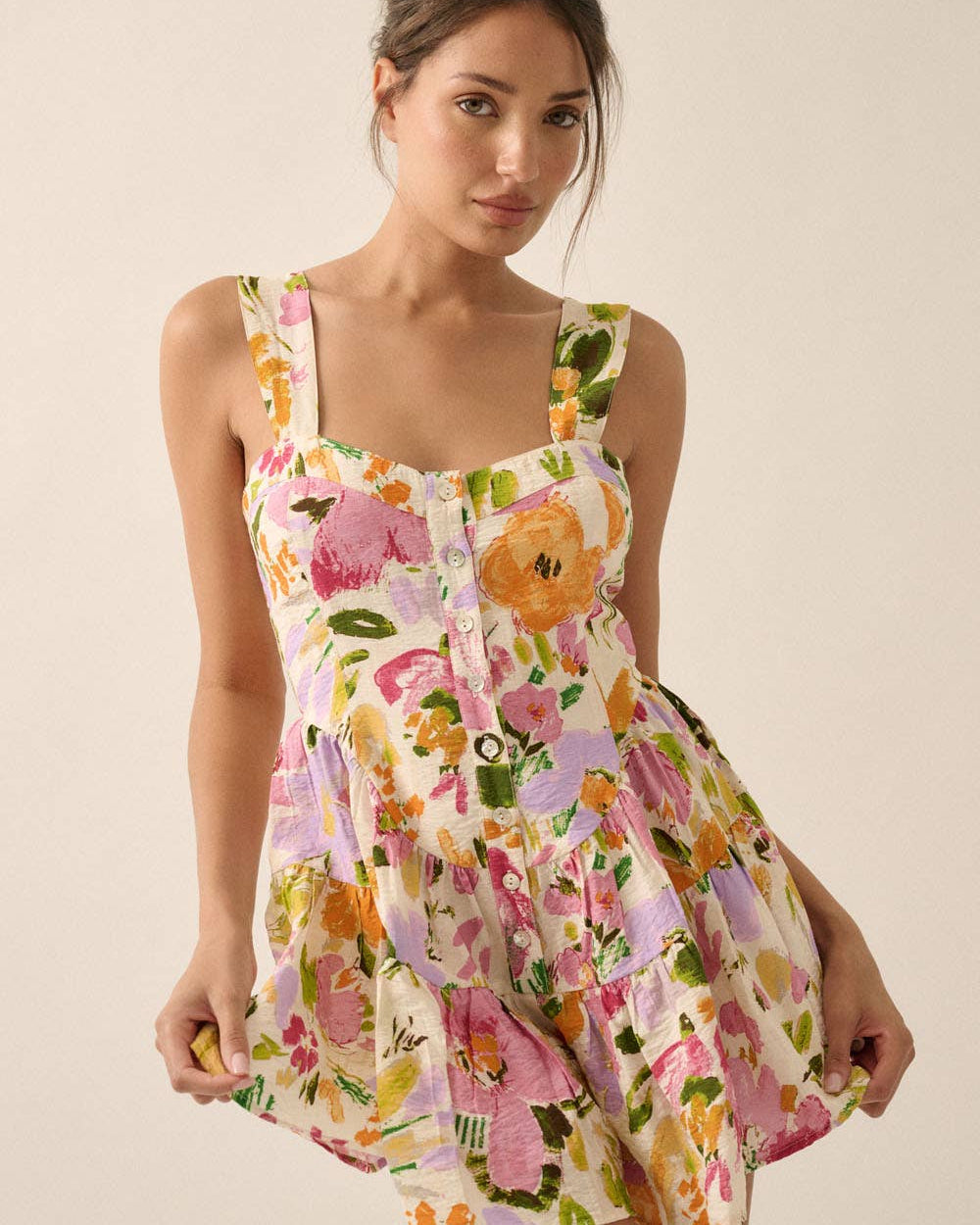 A woman modeling a sleeveless, scoop-neck mini dress with a colorful floral print on a cream background. The dress features a gathered waist and a flared skirt, with a button-down front.