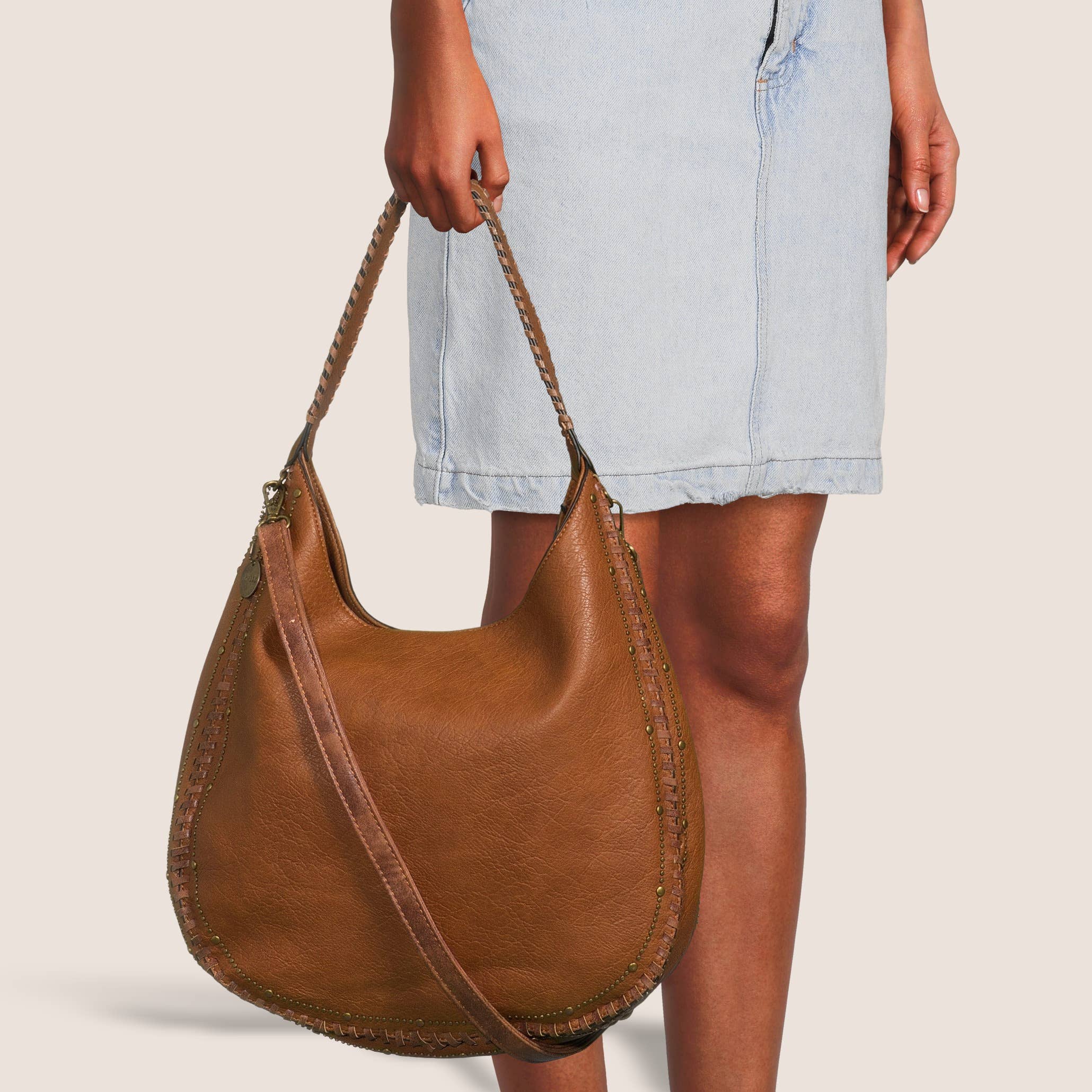 Brown leather handbag held by a person wearing a light blue denim skirt on a beige background
