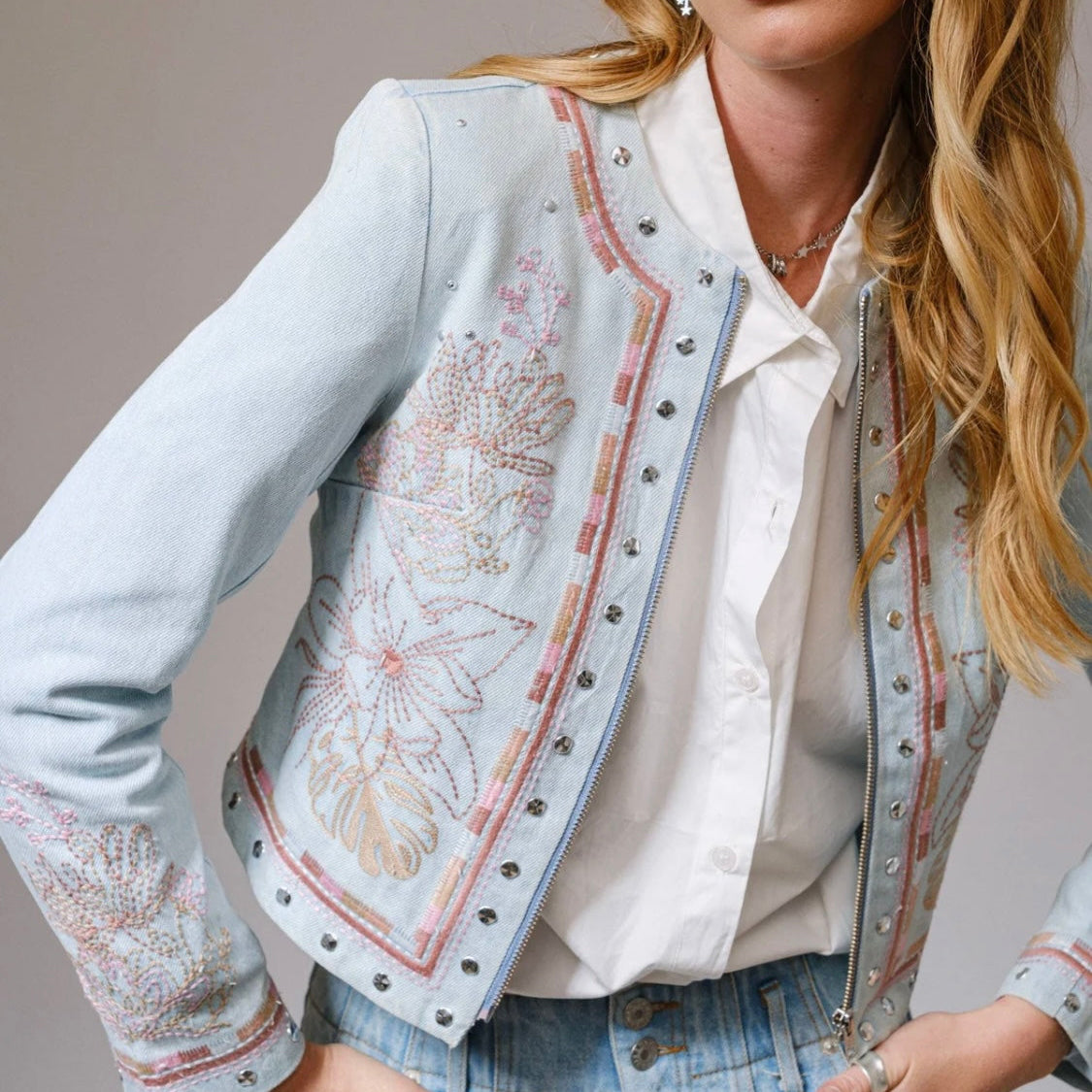 woman wearing light wash embroidered studded denim jacket layered over a white blouse with blue jeans, western boho style detail close up