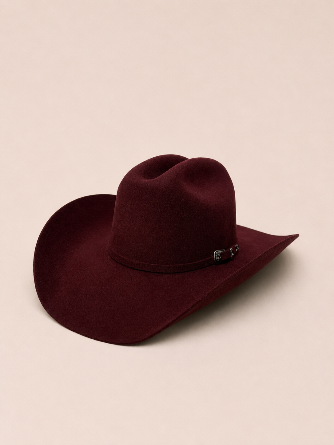 A dark burgundy felt cowboy hat with a matching band and a small decorative black and silver buckle, set against a solid light beige background.